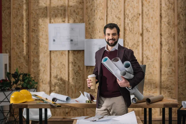 Schöner junger Architekt mit Coffee to go und gerollten Entwürfen am Arbeitsplatz — Stockfoto