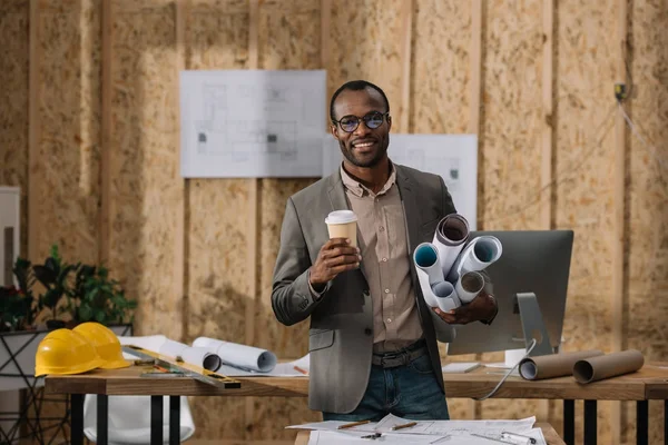 Lächelnder afrikanisch-amerikanischer Architekt mit Coffee to go und gerollten Entwürfen am Arbeitsplatz — Stockfoto