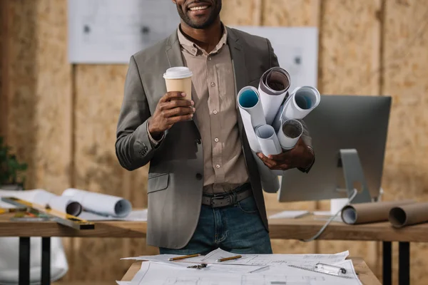 Plan recadré d'architecte souriant avec du café à emporter et plans roulés sur le lieu de travail — Photo de stock