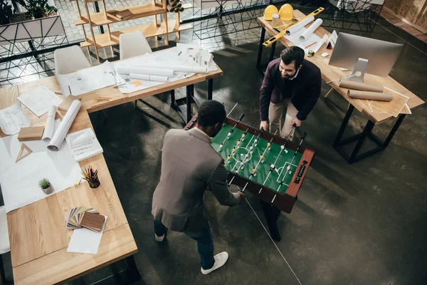 Vue en angle élevé de jeunes architectes jouant au baby-foot dans le bureau plein de plans de construction — Photo de stock