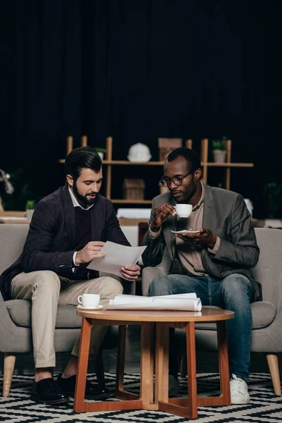 Jeunes hommes d'affaires multiethniques assis dans des fauteuils avec du café et discutant de documents — Photo de stock