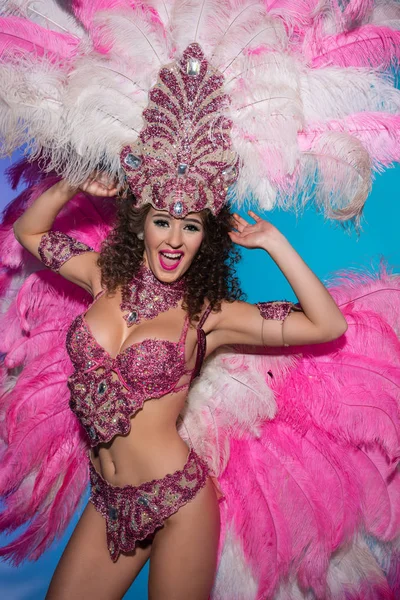 Mujer joven feliz disfrazada de carnaval con plumas rosadas actuando aisladas sobre fondo azul - foto de stock