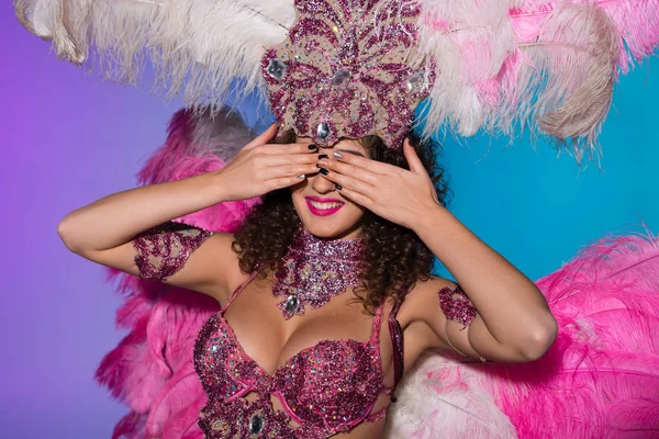 Fröhliche Frau im Karnevalskostüm mit rosafarbenen Federn, die ihre Augen isoliert auf blauem Hintergrund bedecken — Stockfoto