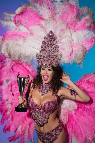 Mujer brillante en traje de carnaval con plumas rosas sosteniendo taza ganadora aislada sobre fondo azul - foto de stock