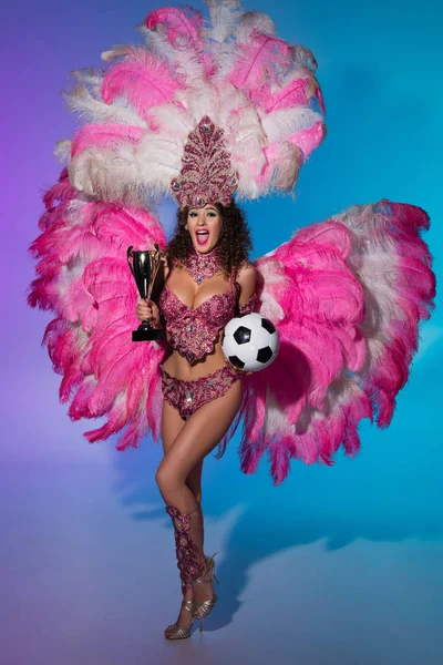 Cheerful woman in carnival costume with pink feathers holding soccer ball and winner cup on blue background — Stock Photo