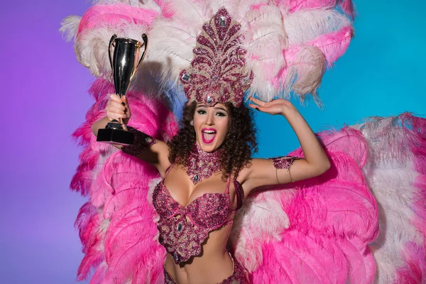 Joyeux jeune femme en costume de carnaval avec des plumes roses tenant tasse gagnante isolé sur fond bleu — Photo de stock