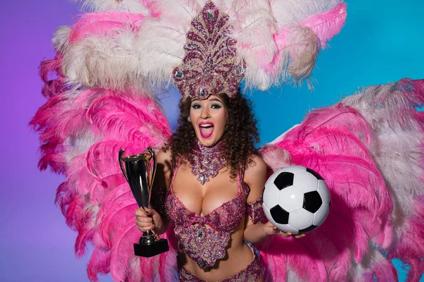 Bright woman in carnival costume with pink feathers holding soccer ball and winner cup on blue background — Stock Photo
