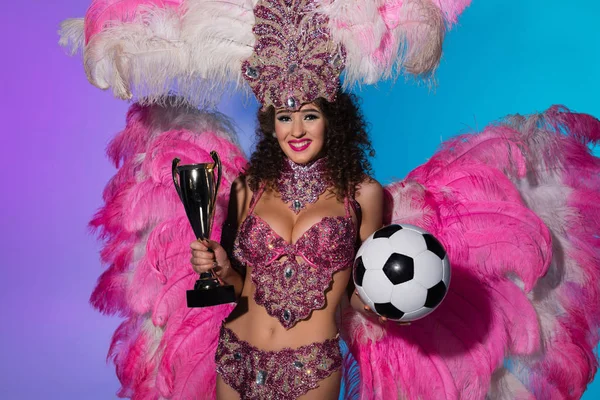 Веселая женщина в карнавальном костюме с розовыми перьями держит футбольный мяч и кубок победителя на синем фоне — стоковое фото