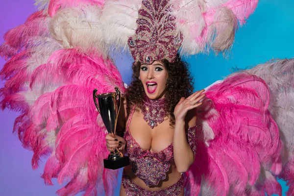 Щаслива молода жінка в карнавальному костюмі з рожевим пір'ям, що тримає кубок переможця ізольовано на синьому фоні — стокове фото