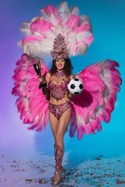 Happy young woman in carnival costume with pink feathers holding soccer ball and winner cup on blue background — Stock Photo