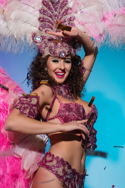 Mujer brillante en traje de carnaval con plumas rosas bailando y sonriendo sobre fondo azul - foto de stock