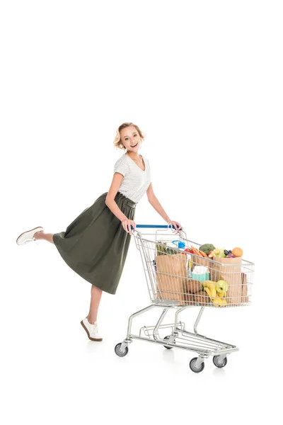 Счастливая молодая женщина, стоящая с тележкой для покупок и улыбающаяся на камеру, изолированную на белом — стоковое фото
