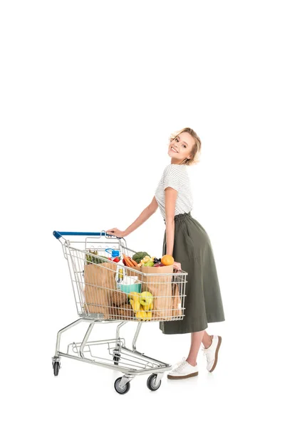 Cheerful young woman standing with shopping trolley and smiling at camera isolated on white — Stock Photo