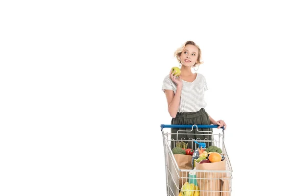 Улыбающаяся молодая женщина, держащая яблоко и глядя в сторону, стоя с тележкой с продуктовыми пакетами, изолированными на белом — стоковое фото