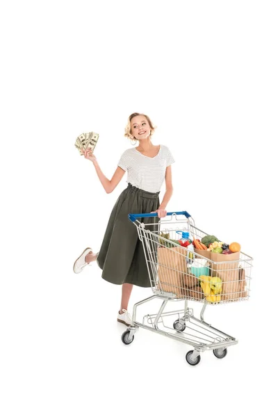 Glückliche junge Frau hält Dollarnoten in der Hand und steht mit Einkaufswagen voller Lebensmittel isoliert auf weißem Grund — Stockfoto