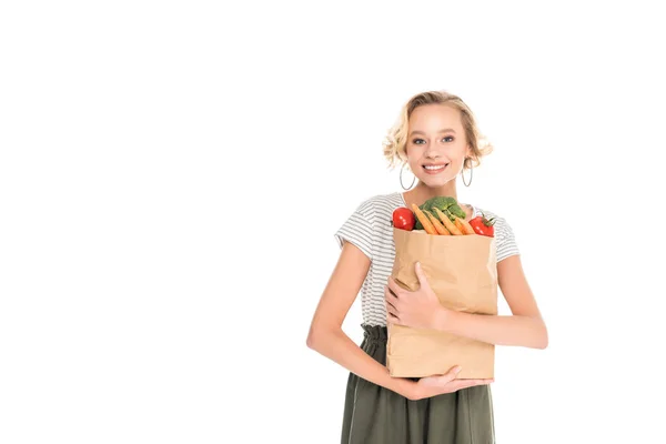 Atractiva mujer joven feliz sosteniendo bolsa de comestibles y mirando a la cámara aislada en blanco - foto de stock