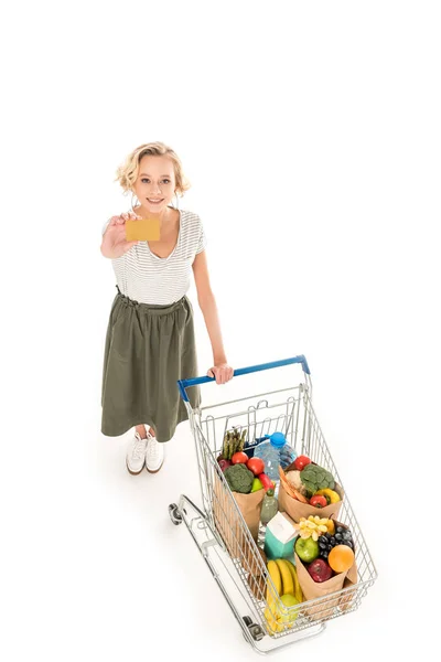 Hochwinkelaufnahme einer jungen Frau, die ihre Kreditkarte hält und in die Kamera lächelt, während sie mit einem Einkaufswagen voller Lebensmittel isoliert auf weiß steht — Stockfoto