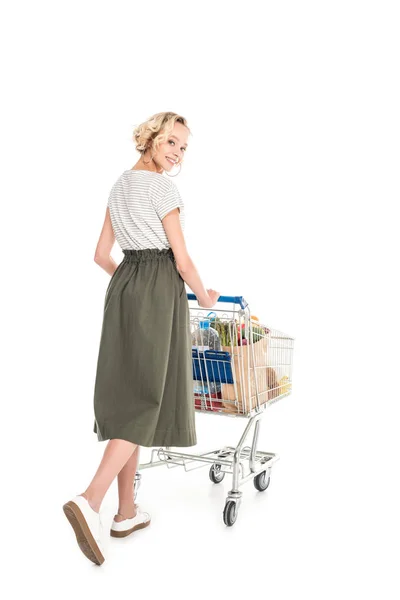 Hermosa mujer joven sonriendo a la cámara mientras empuja el carro de la compra con bolsas de comestibles aisladas en blanco - foto de stock