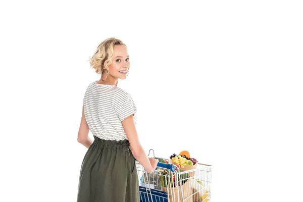 Hermosa chica sonriendo a la cámara mientras empuja el carro de la compra con bolsas de comestibles y botella de plástico de agua aislada en blanco - foto de stock