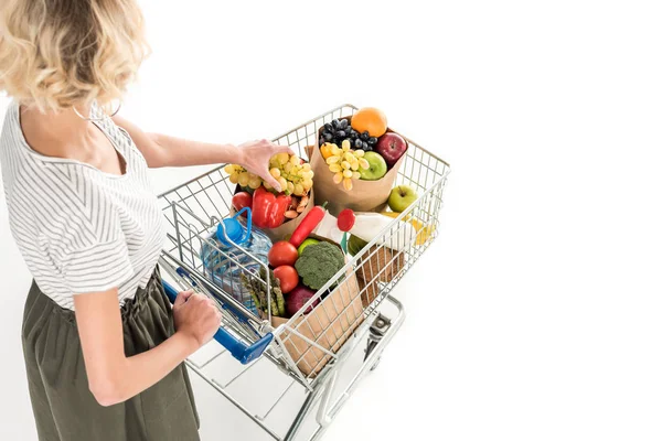 Cropped shot of young woman pushing shopping trolley with grocery bags and plastic bottle of water isolated on white — Stock Photo
