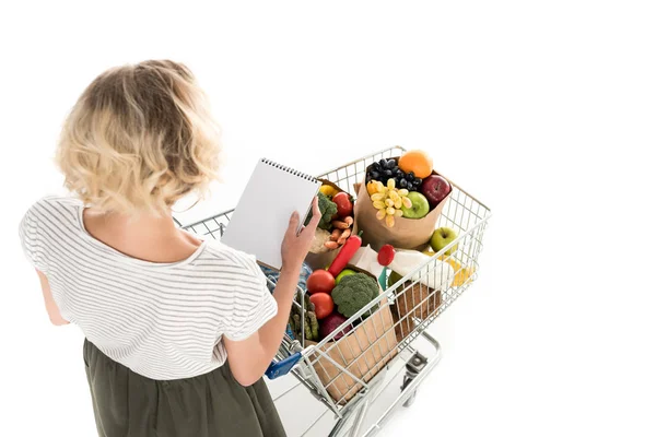 Visão de alto ângulo da jovem segurando caderno em branco e carrinho de compras com sacos de supermercado isolados em branco — Fotografia de Stock