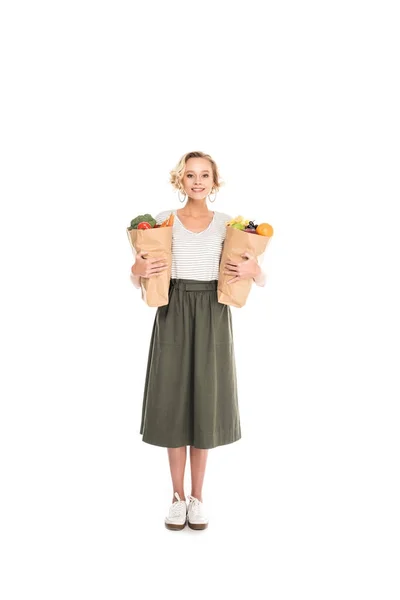 Vue pleine longueur de la femme tenant des sacs en papier avec des fruits et légumes et souriant à la caméra isolée sur blanc — Photo de stock