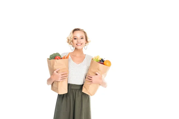 Young woman holding paper bags with fruits and vegetables and smiling at camera isolated on white — Stock Photo