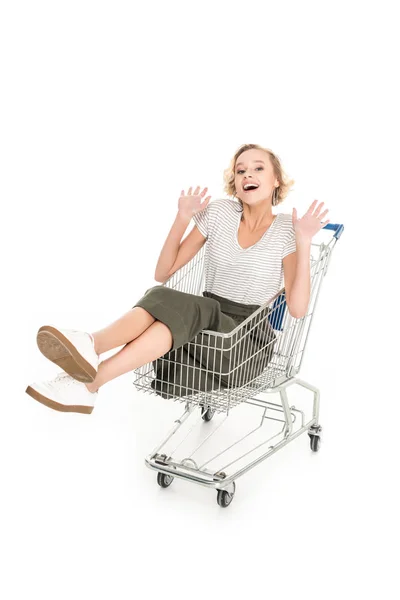 Cheerful young woman sitting in shopping trolley and smiling at camera isolated on white — Stock Photo