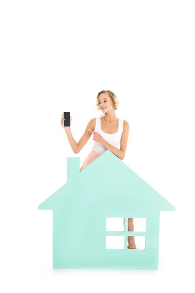 Young woman pointing at smartphone in hand while standing near house model isolated on white — Stock Photo