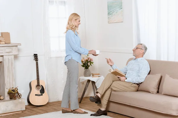 Älterer Mann liest Buch, während Frau ihm Tee im gemütlichen Zimmer serviert — Stockfoto