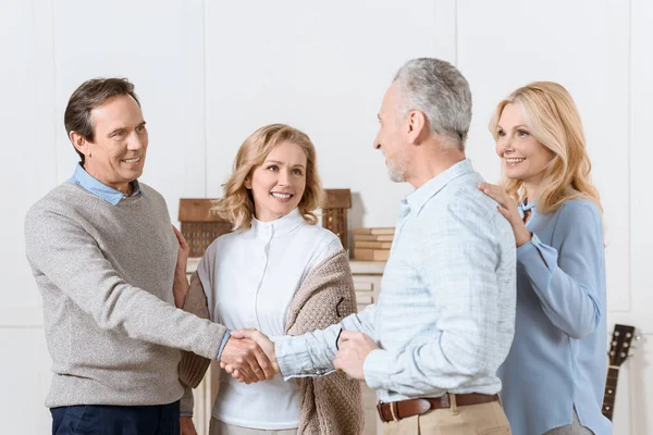 Pareja de hombres y mujeres de mediana edad saludando a sus amigos como invitados y dándoles la mano - foto de stock