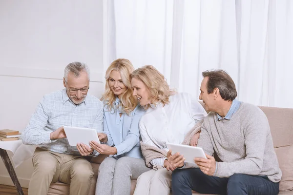 Hommes et femmes d'âge moyen utilisant des tablettes numériques assis sur le canapé — Photo de stock