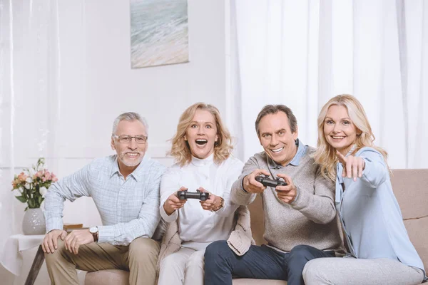 Hombres y mujeres de mediana edad sentados en el sofá y jugando a la consola con joysticks - foto de stock