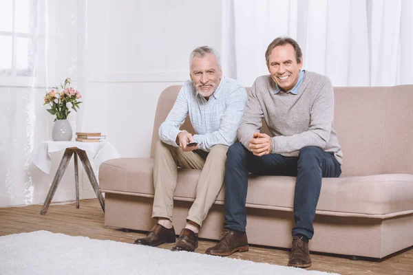 Dos hombres adultos felices sentados en el sofá en la sala de estar ligera y viendo la televisión - foto de stock