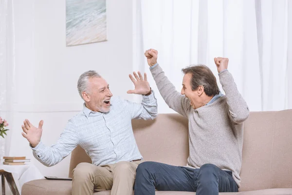 Two happy adult men sitting on sofa and celebrating elated in light living room — Stock Photo