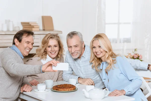 Uomo facendo selfie mentre gli amici seduti a tavola con tè e torta — Foto stock