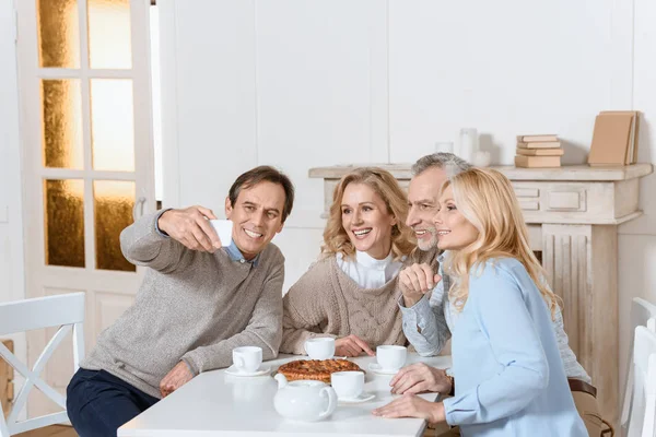 Hombre haciendo selfie mientras amigos sentado en la mesa con té y pastel - foto de stock