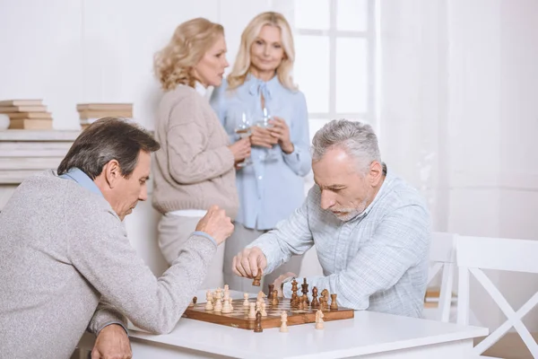 Hommes jouant aux échecs assis à table, femme debout avec des lunettes sur le fond — Photo de stock