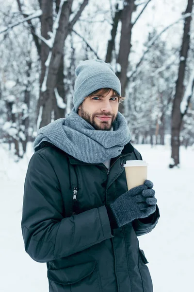 Portrait de beau jeune homme avec café pour aller dans les mains dans un parc enneigé — Photo de stock