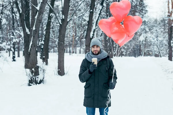 Retrato de hombre joven con café para llevar y globos en forma de corazón en el parque de invierno - foto de stock