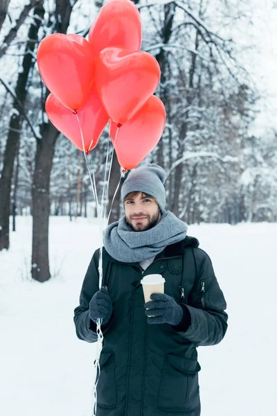 Портрет молодого человека с кофе и воздушными шарами в форме сердца в зимнем парке — стоковое фото