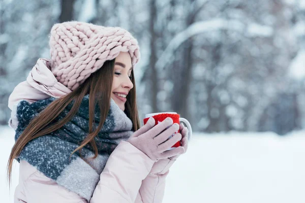 Vista lateral de la hermosa mujer joven con taza de café caliente en las manos en el parque cubierto de nieve - foto de stock