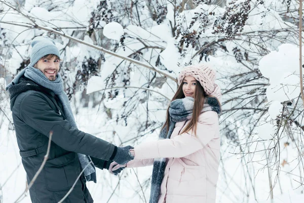 Sonriente pareja cogida de la mano y mirando a la cámara en el bosque nevado - foto de stock