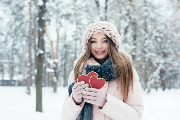 Portrait of beautiful young woman with hearts in hands looking at camera in snowy park — Stock Photo