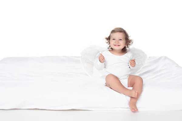 Adorabile bambino felice con le ali seduto sul letto, isolato su bianco — Foto stock