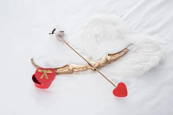 Vue du dessus de la boîte cadeau, ailes, arc et flèche sur lit blanc — Photo de stock