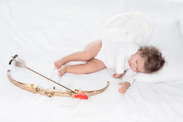 Little angel with wings lying on bed with bow and arrow — Stock Photo