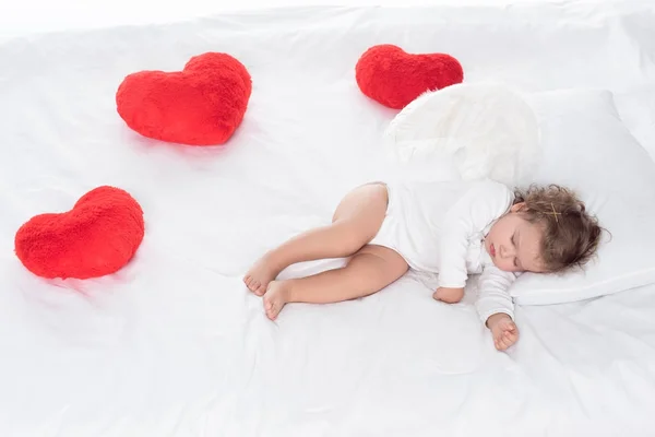 Little angel with wings lying on bed with hearts — Stock Photo