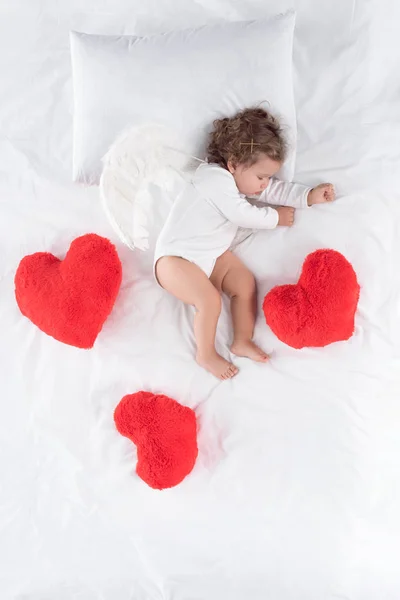 Little cupid with wings lying on bed with red hearts — Stock Photo