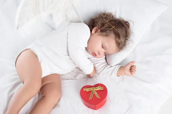 Kleiner Cherub mit Flügeln auf Bett liegend mit Geschenkbox — Stockfoto